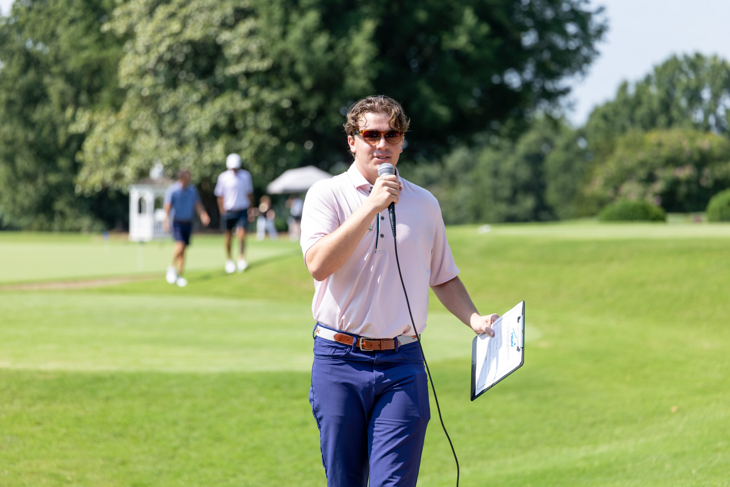 A CCVA Golf Pro holding a microphone and clipboard stand on a golf course during an event.