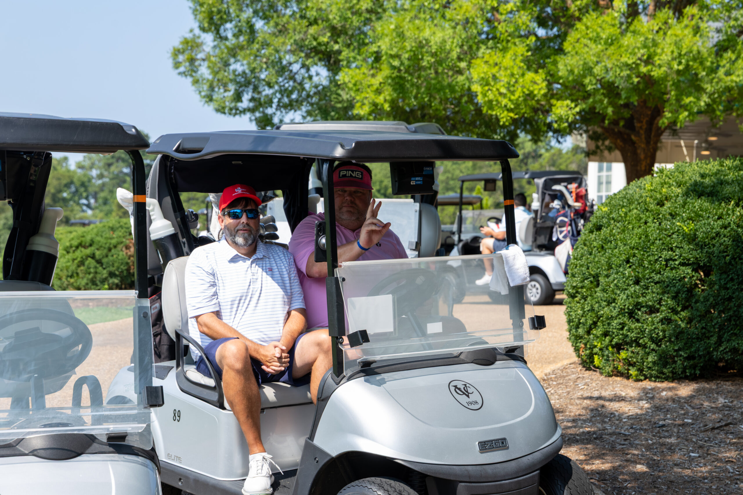 Two men sitting on a golf cart in front of green trees and bushes.