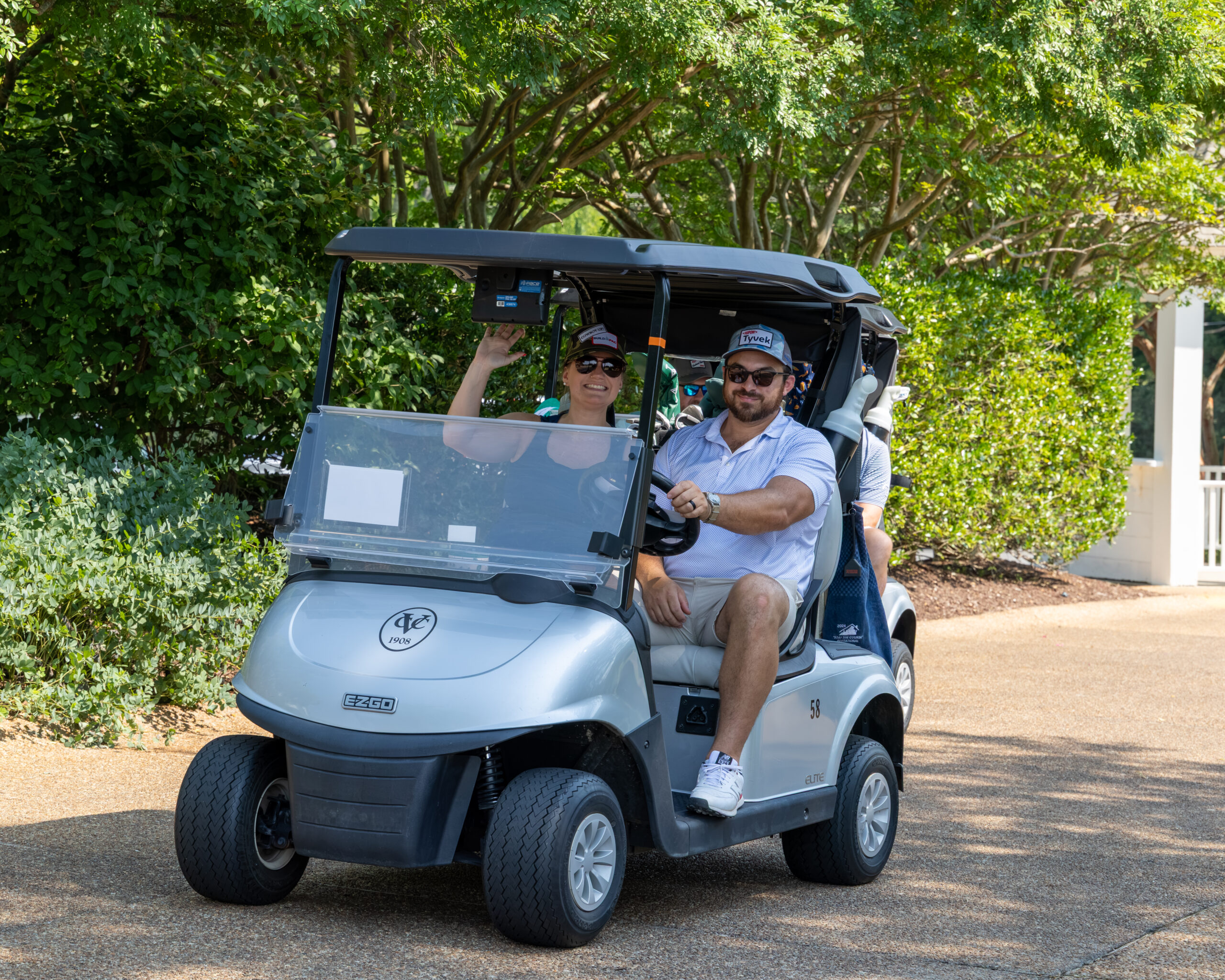 Two golfers sitting in a golf cart on a paved path near trees and shrubbery.