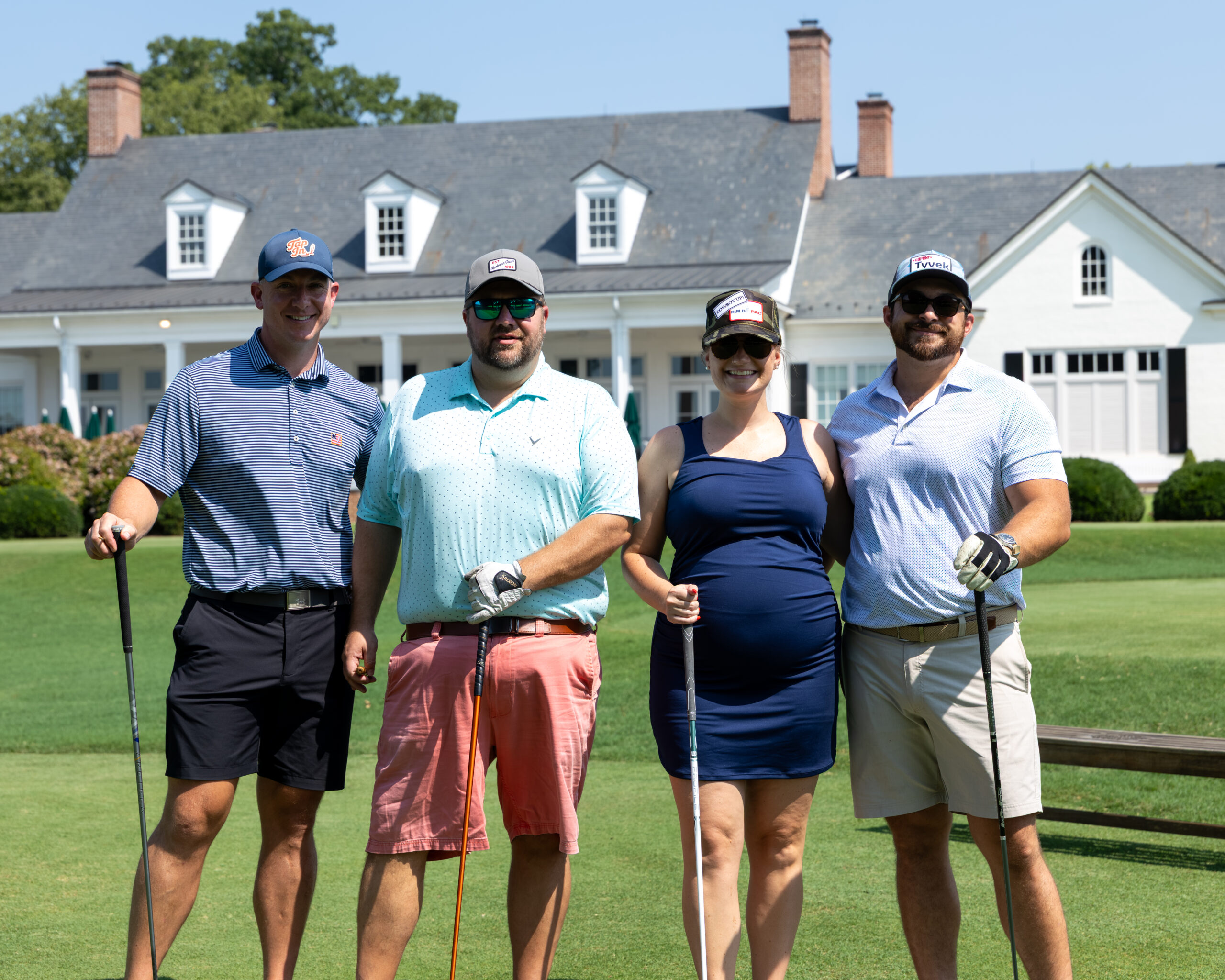 Four people posing with golf clubs in front of a clubhouse building.