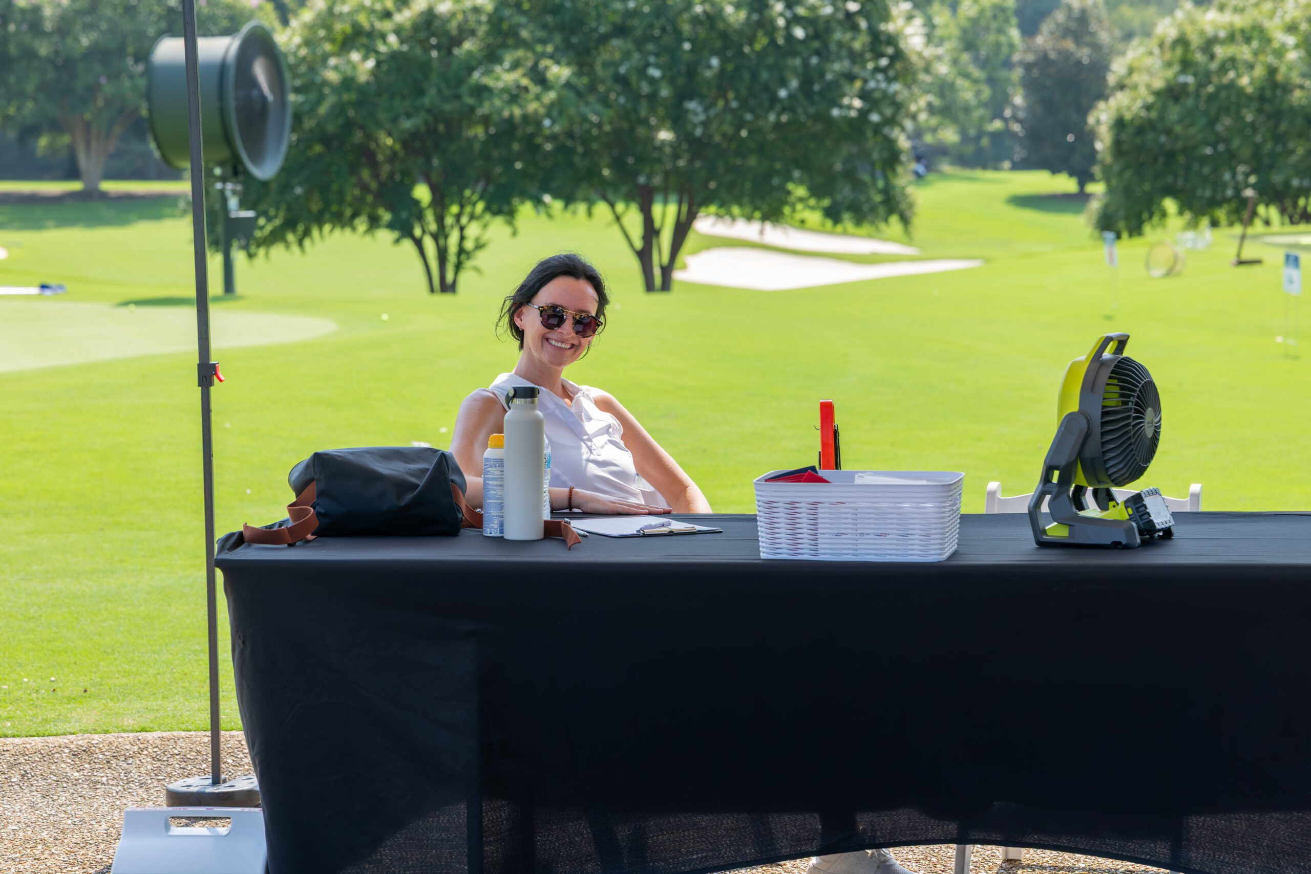 A woman with sunglasses smiling at a table outdoors on a golf course.