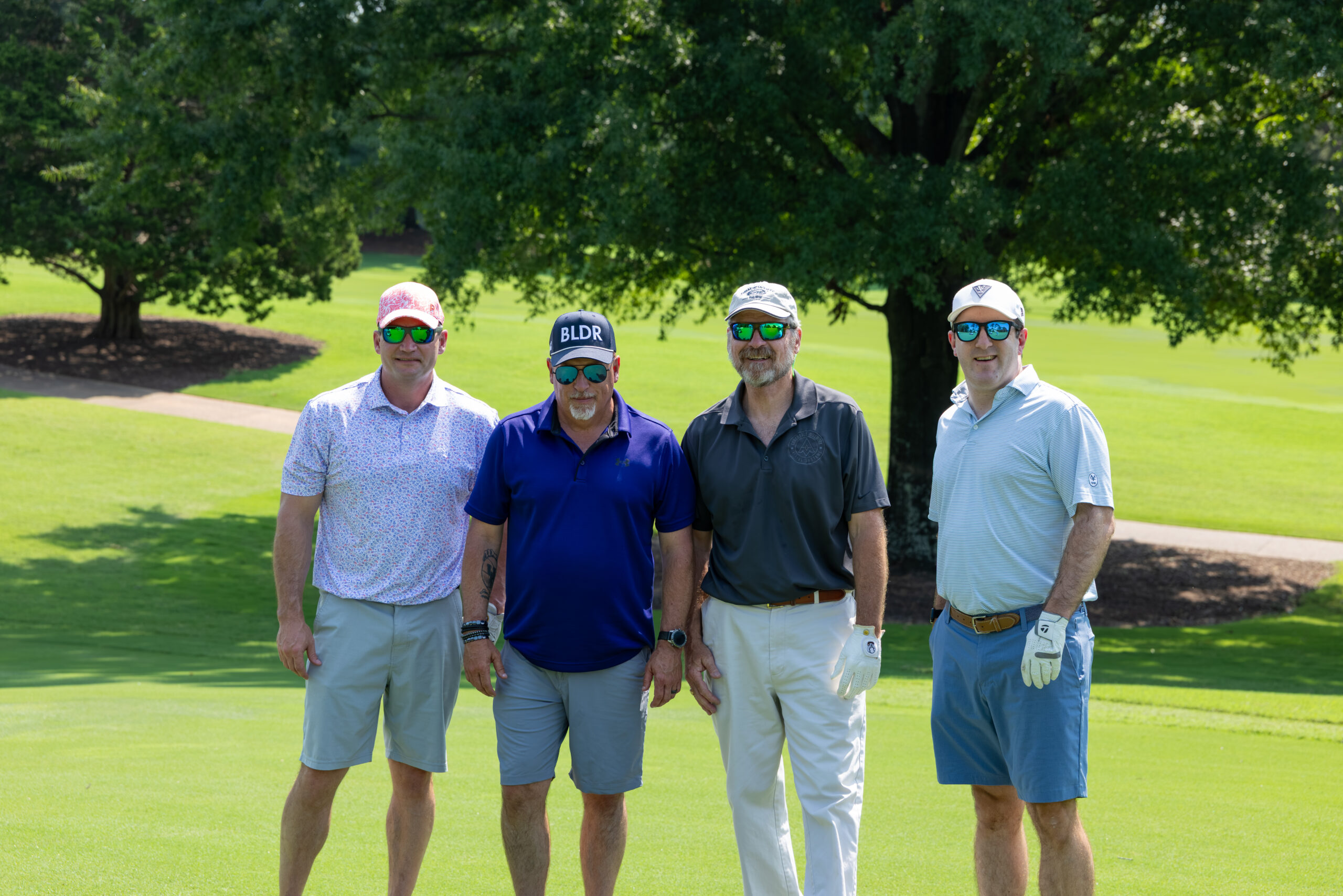 Four men in golf attire standing on a green grass field near trees.