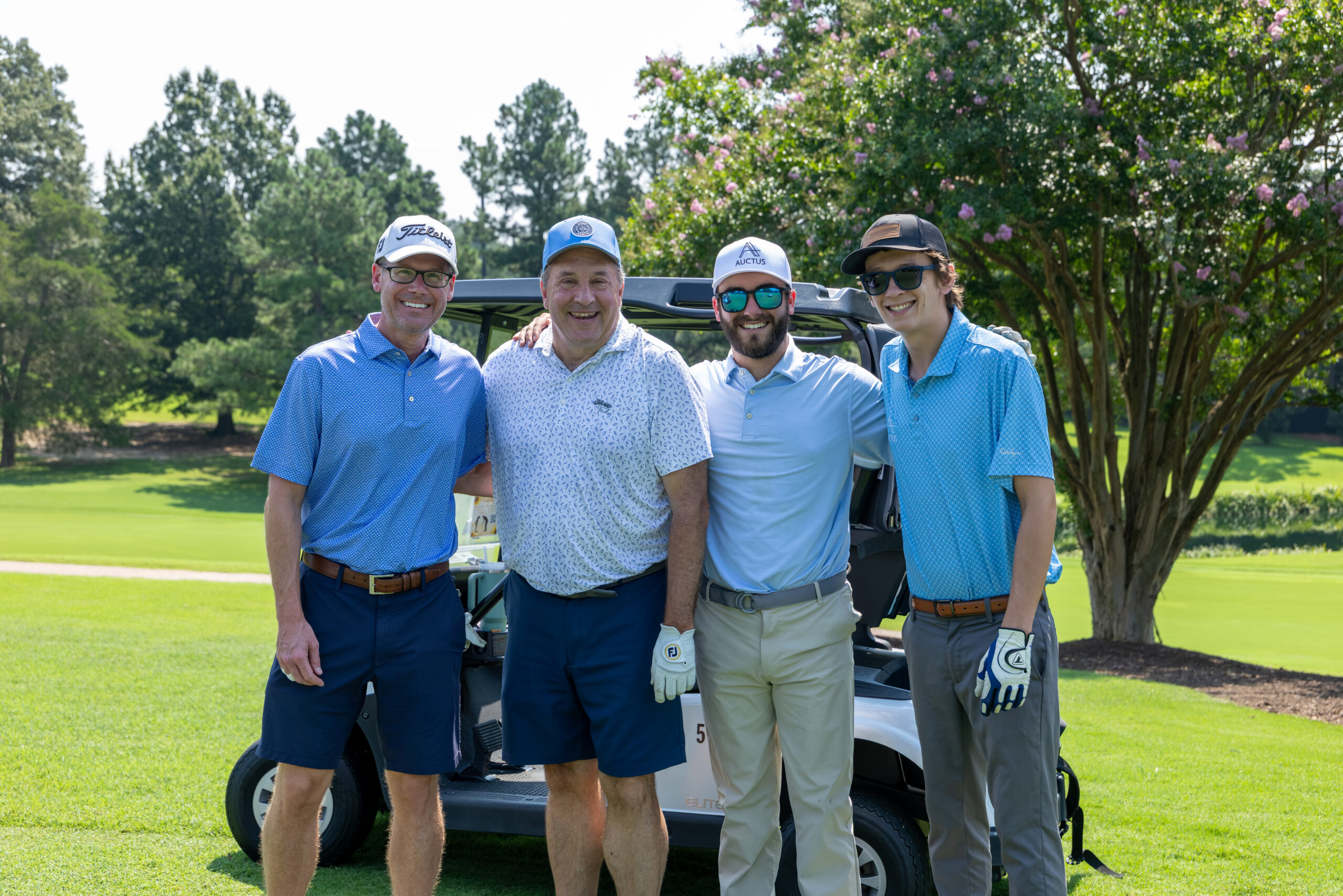Four men in golf attire standing together in front of a golf cart on a sunny course.