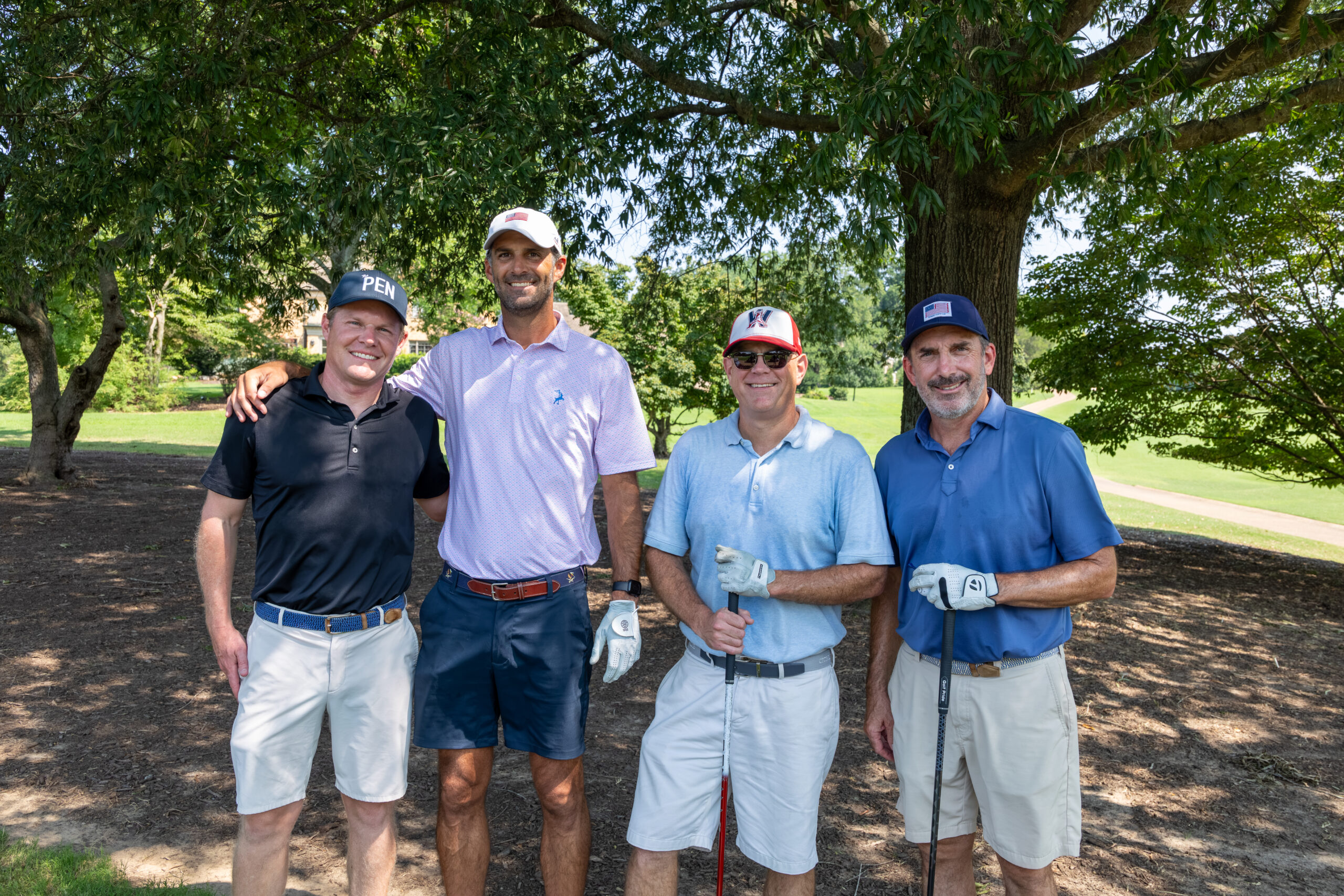 Four men in golf attire standing together under a large tree on a golf course.