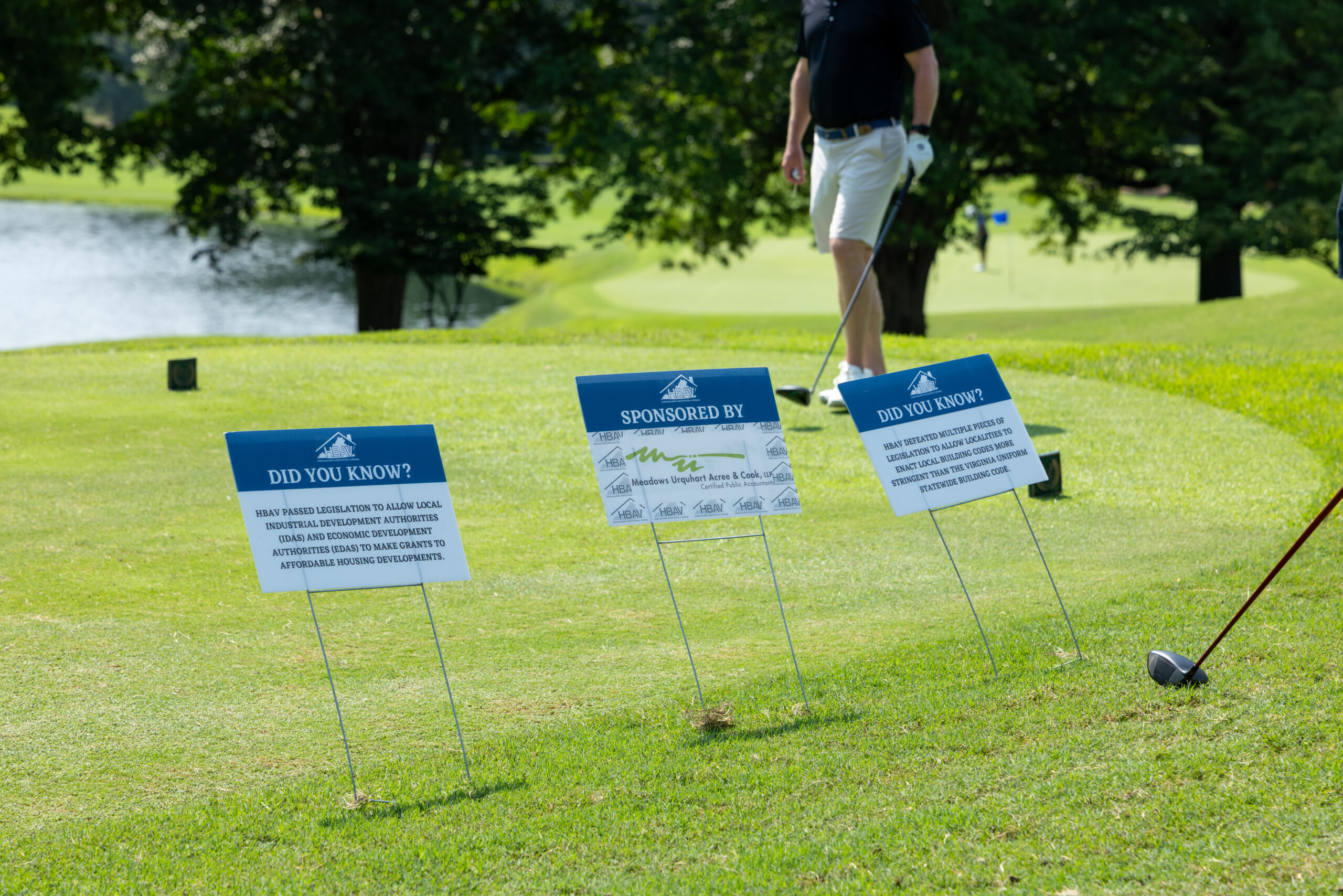 Three golf tee signs with sponsorship logos and facts, with a golfer walking in the background on a sunny day.