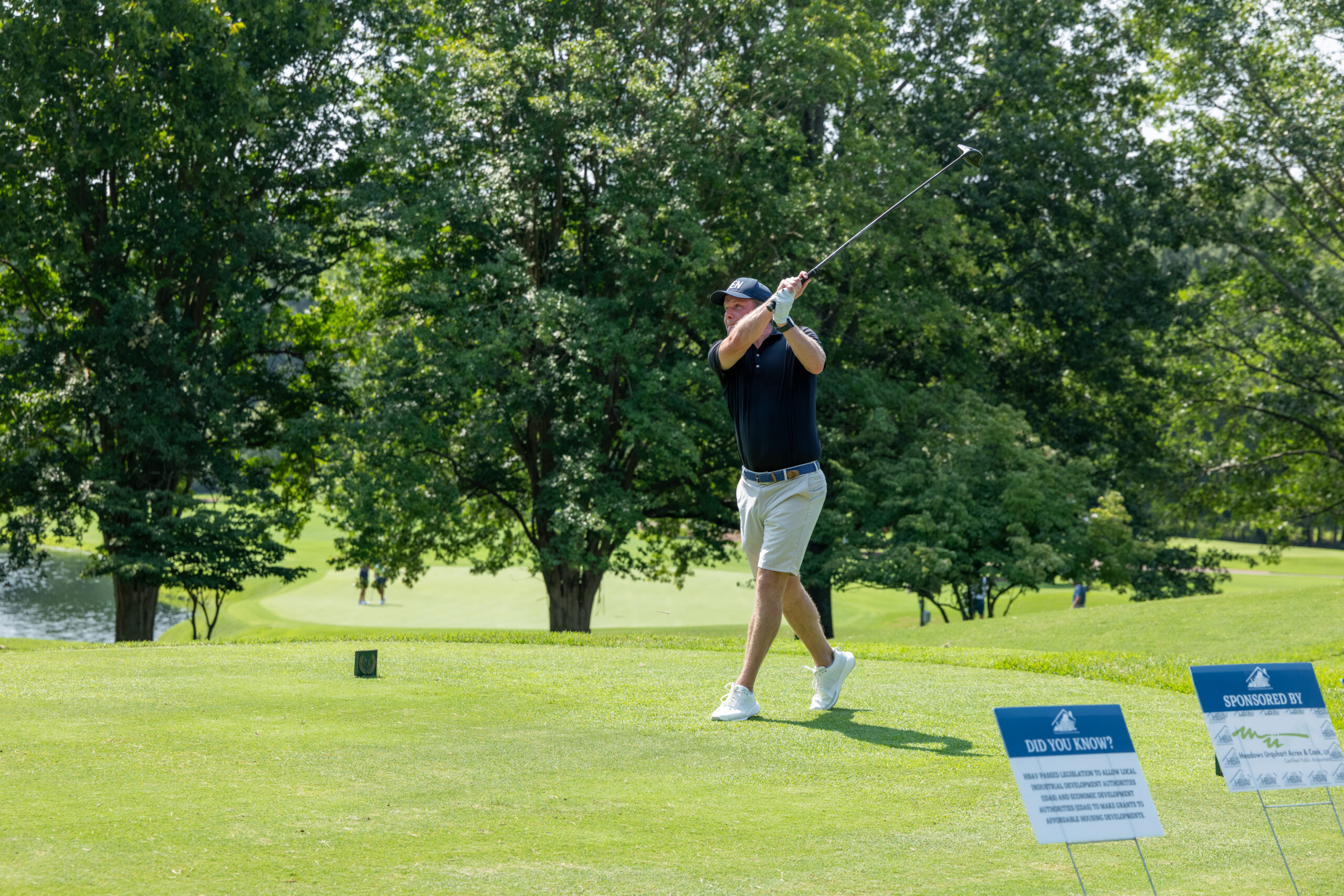 A golfer takes a swing with a club during a tournament on a grassy golf course near trees and water.