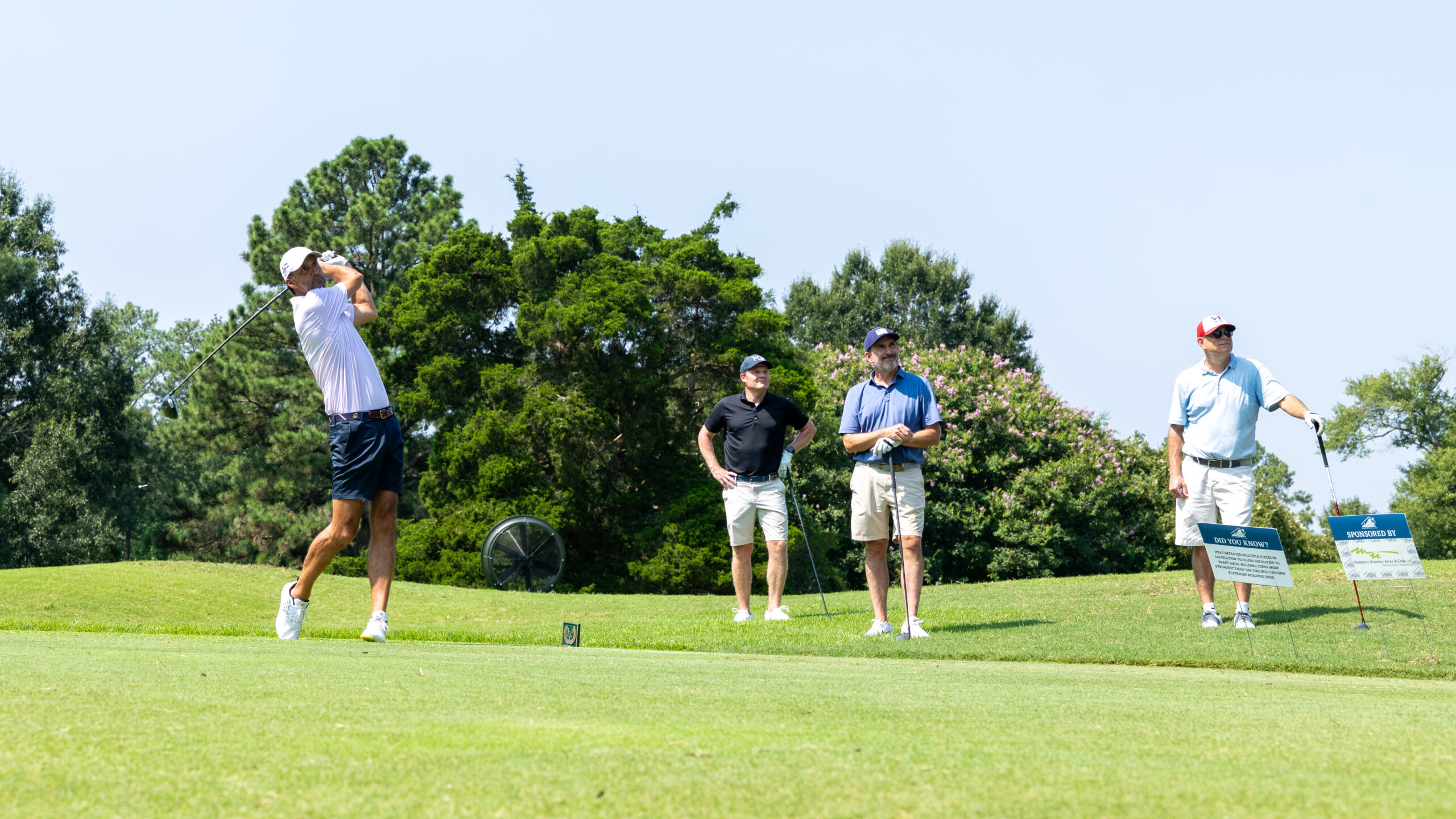 Three men golfers playing on a green field with trees and bushes in background.