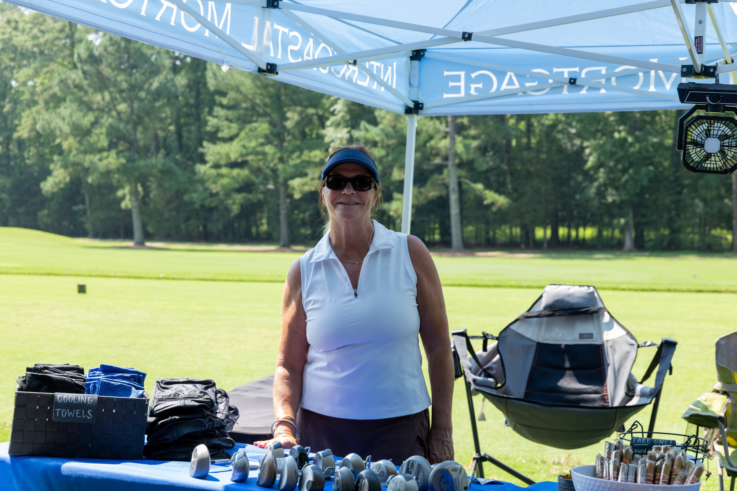 A woman in a white sleeveless shirt and visor stands behind a table at an outdoor golf event.