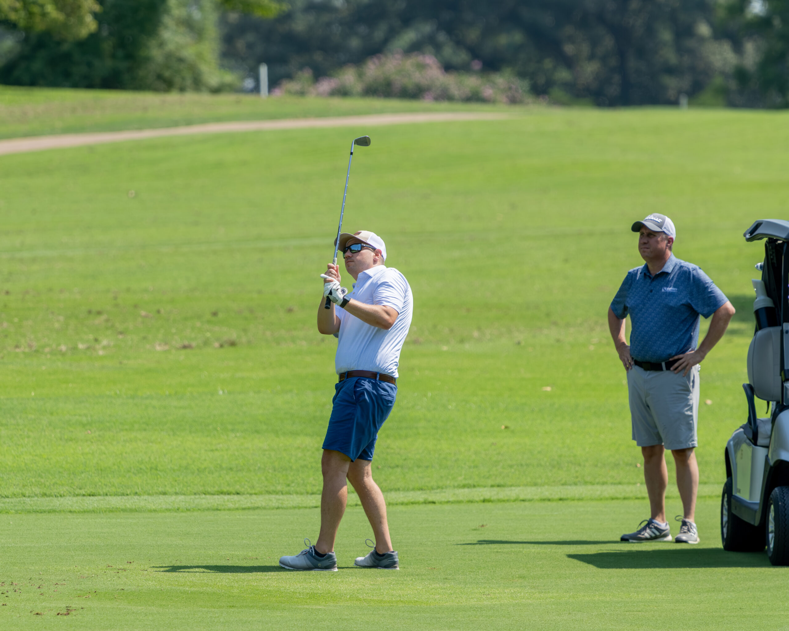 A man in a white polo shirt and blue shorts is practicing his golf swing on a green fairway.