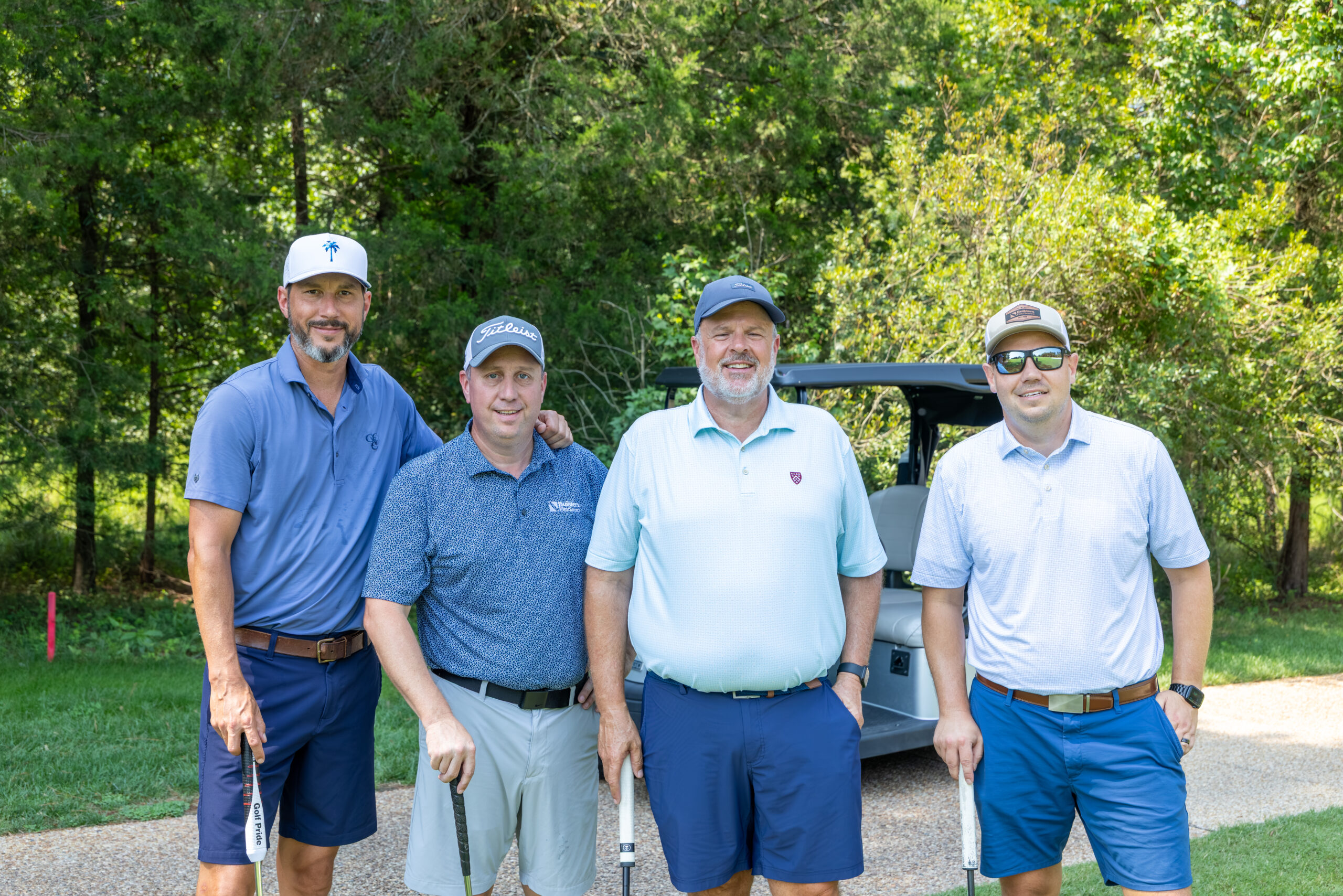 Four men in golf attire standing in front of a golf cart near a wooded area.