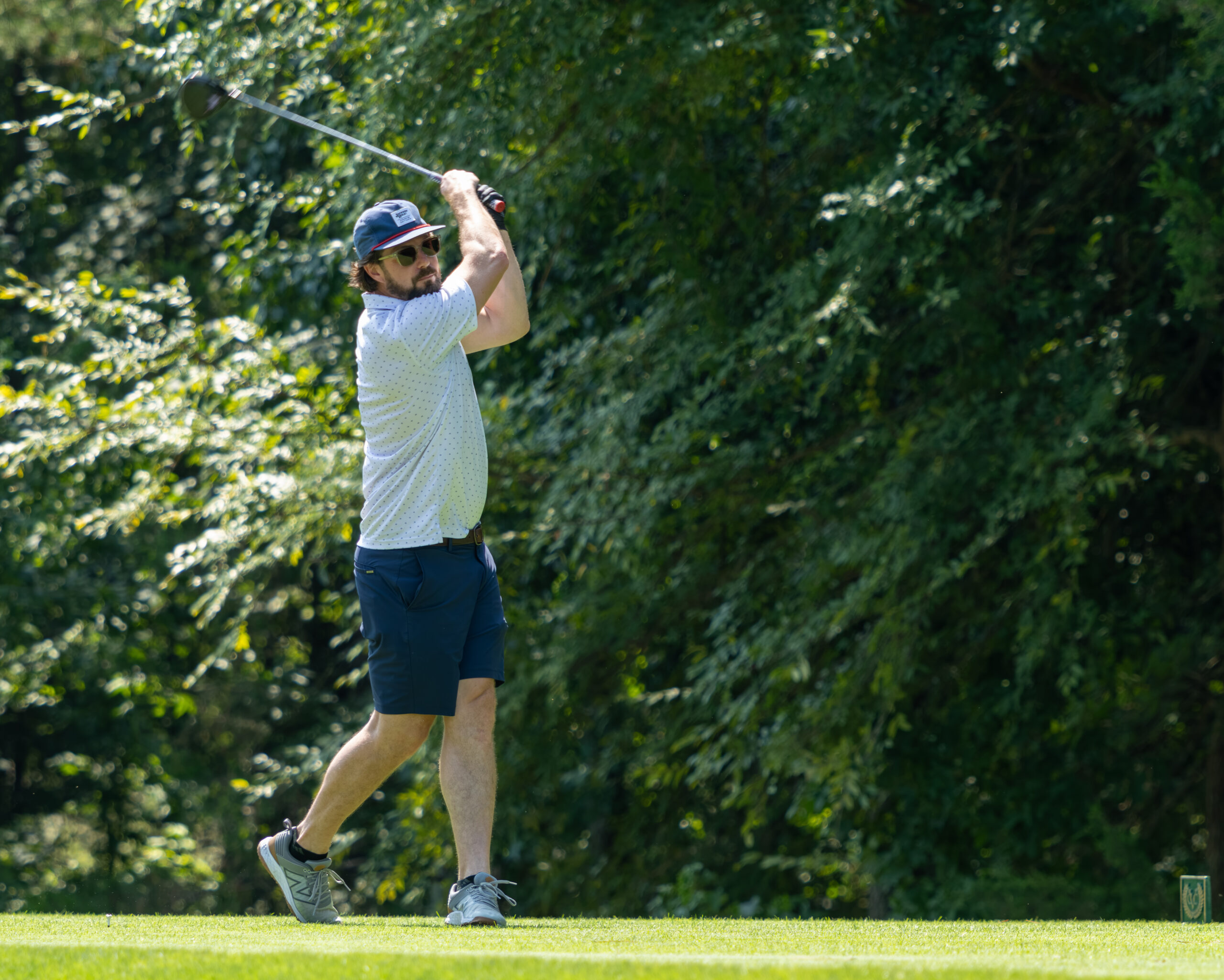 A man in a polo shirt and shorts golfs near trees.