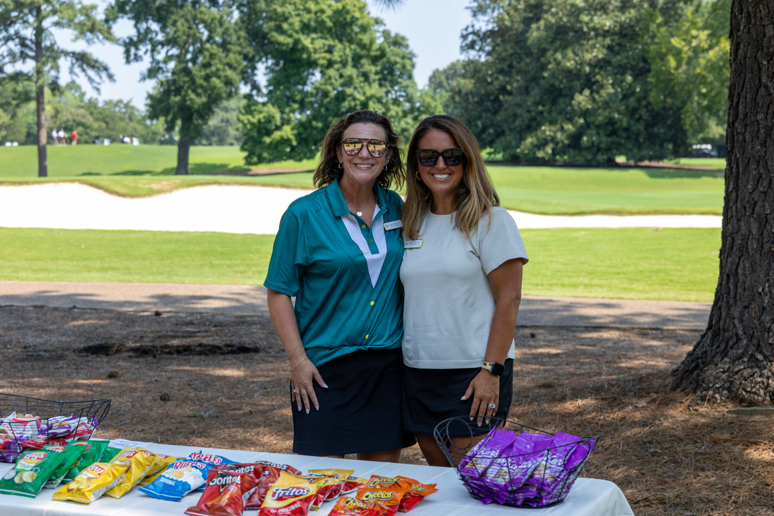 Two women standing together on a golf course behind a table of bagged snacks and products.