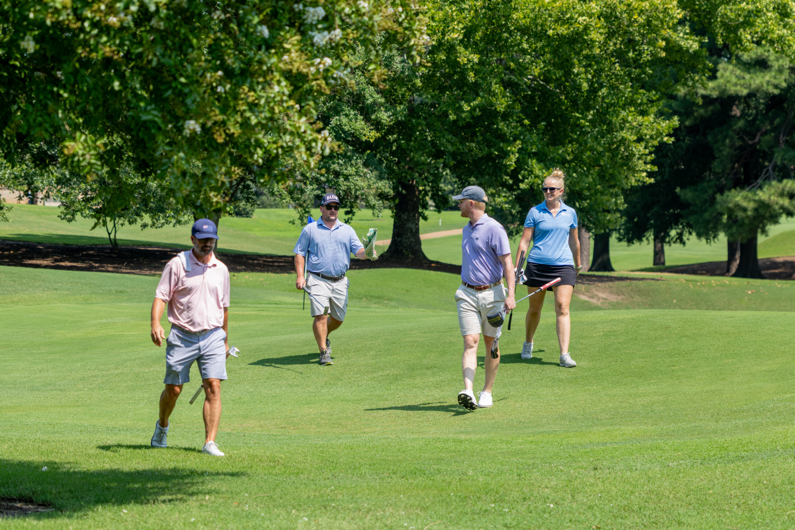 Four golfers dressed in casual attire playing golf on a grassy green field among trees.