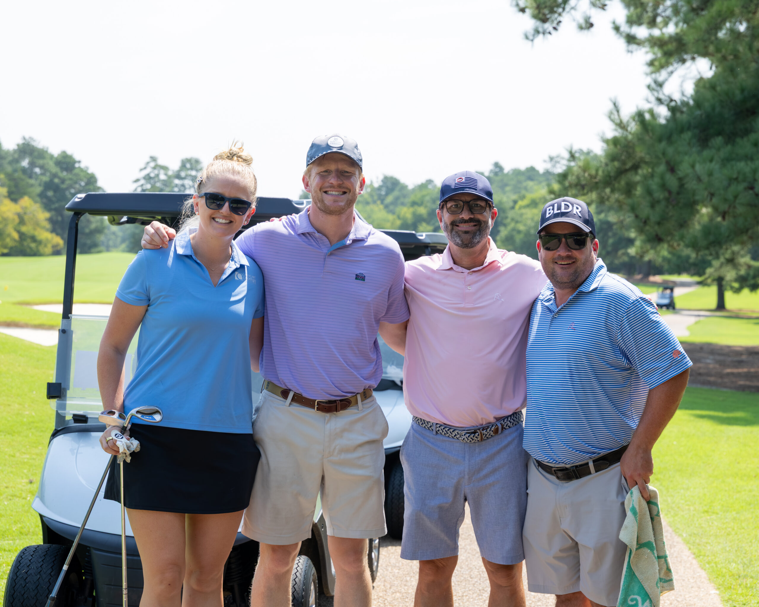 Four people pose for a photo in front of a golf cart on a green course.