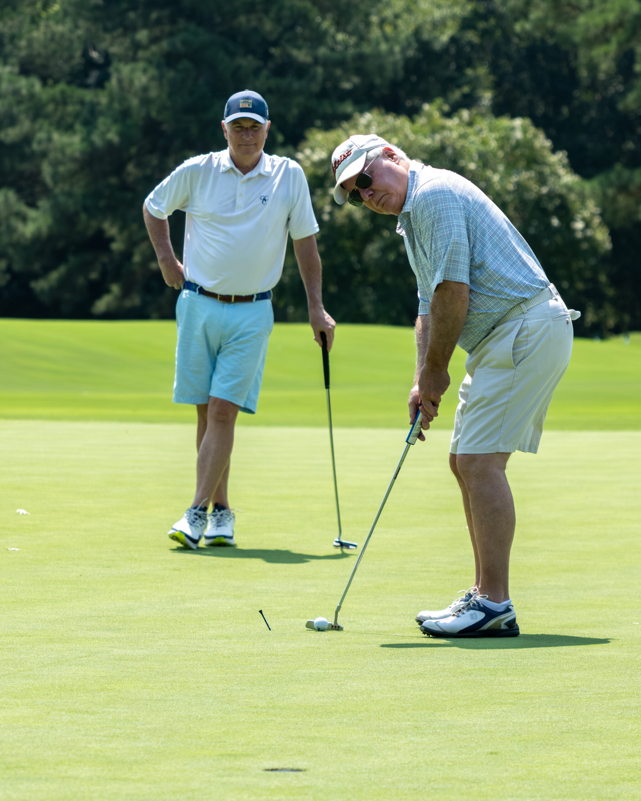 Two men on a golf course, one leaning over his club to line up a shot with another man watching.