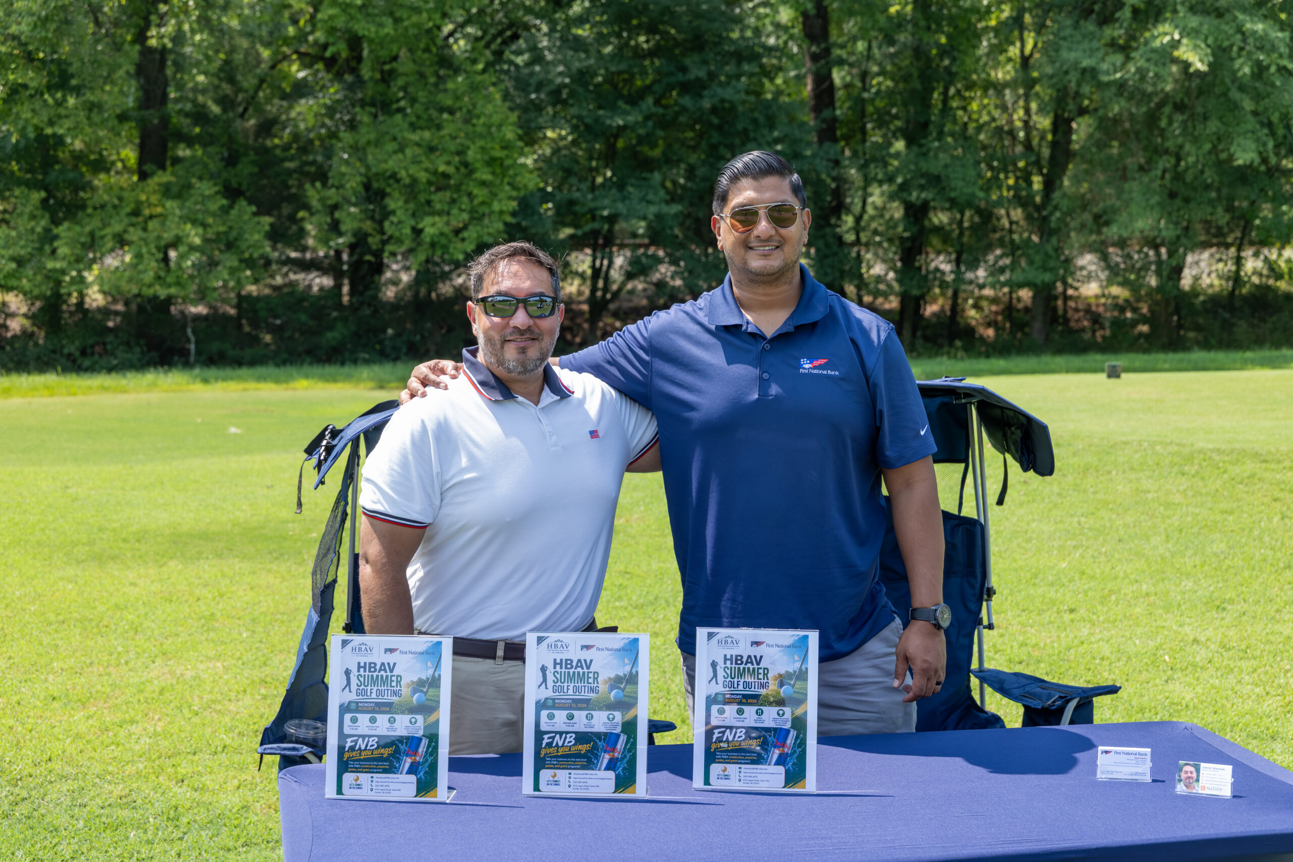 Two men standing behind a table with flyers and a banner on a golf course.