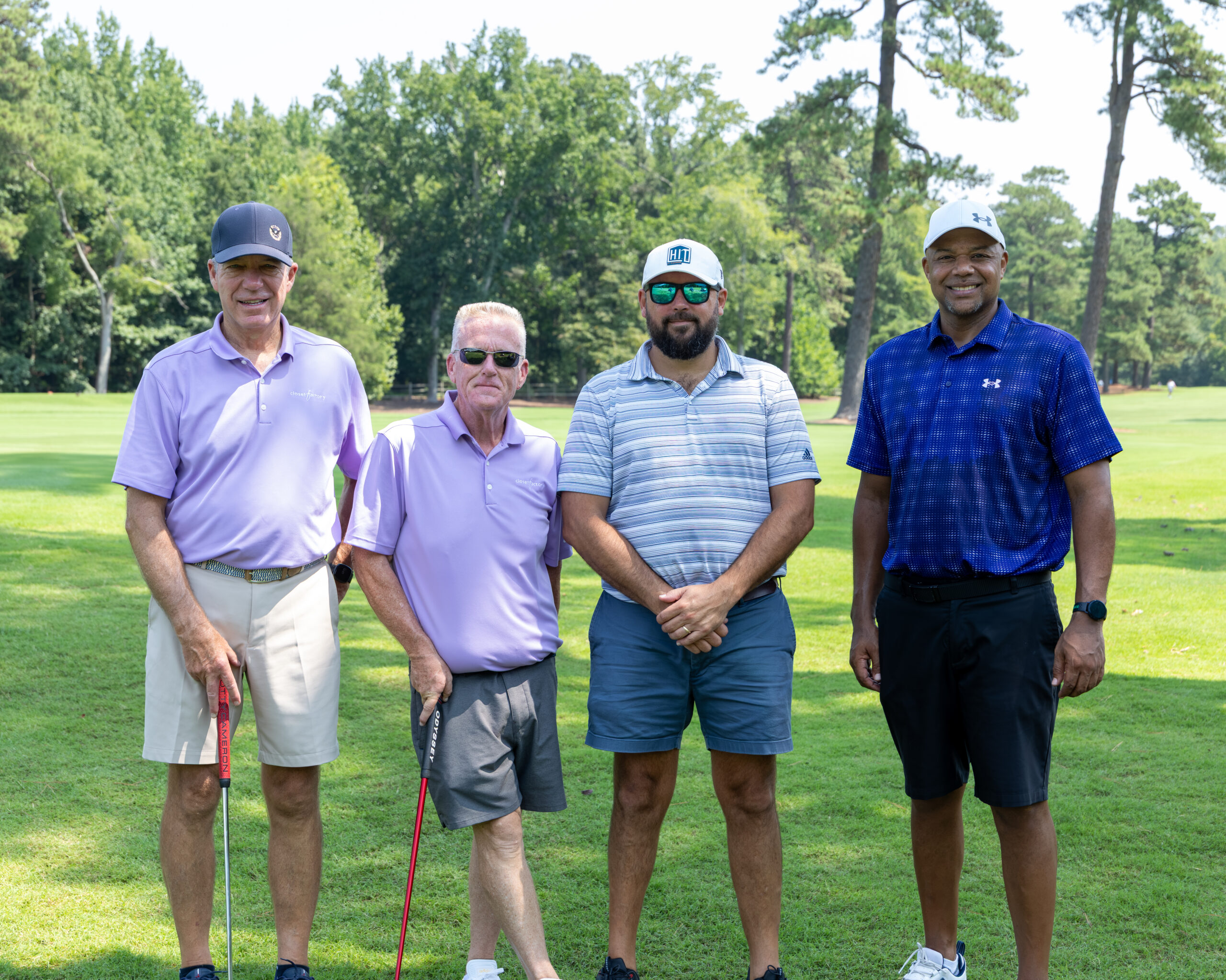 Four men in golf attire standing together on a grassy field with trees and woods in the background.