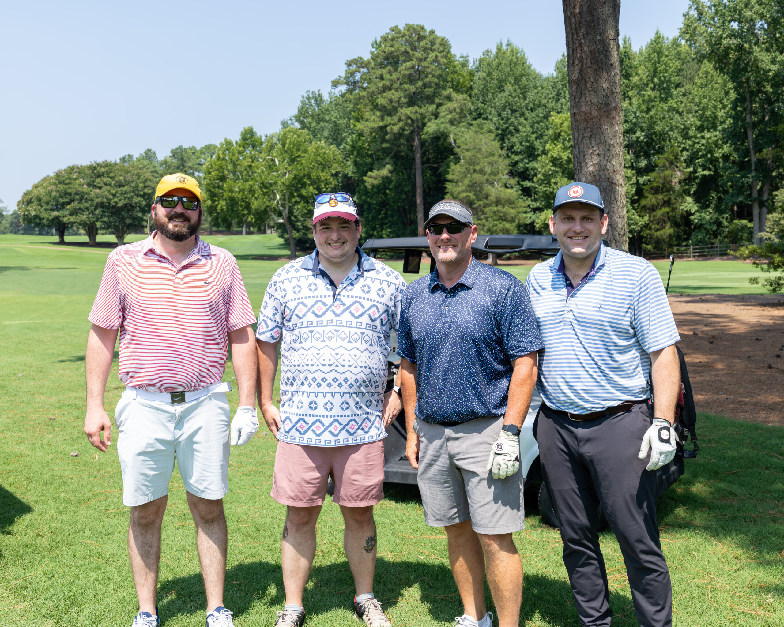 Four men standing together in golf attire on a grassy field near trees.