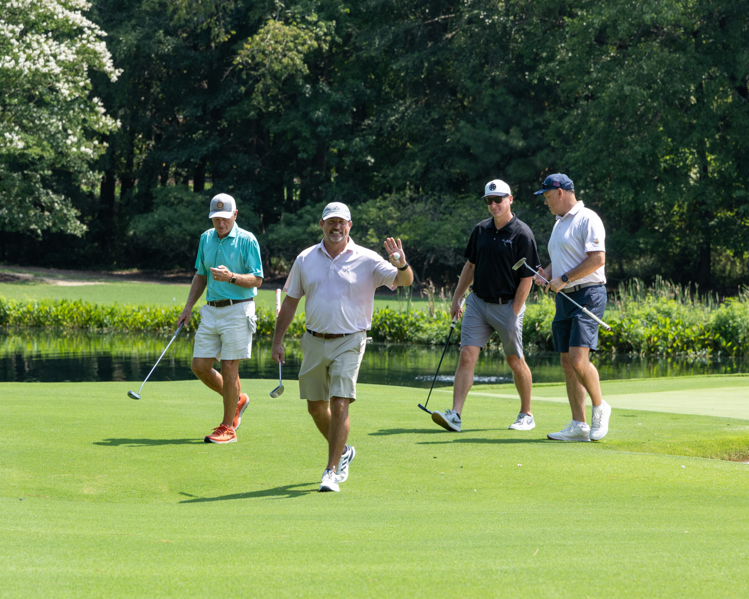 Four men playing golf and posing on a green near water with trees in the background.