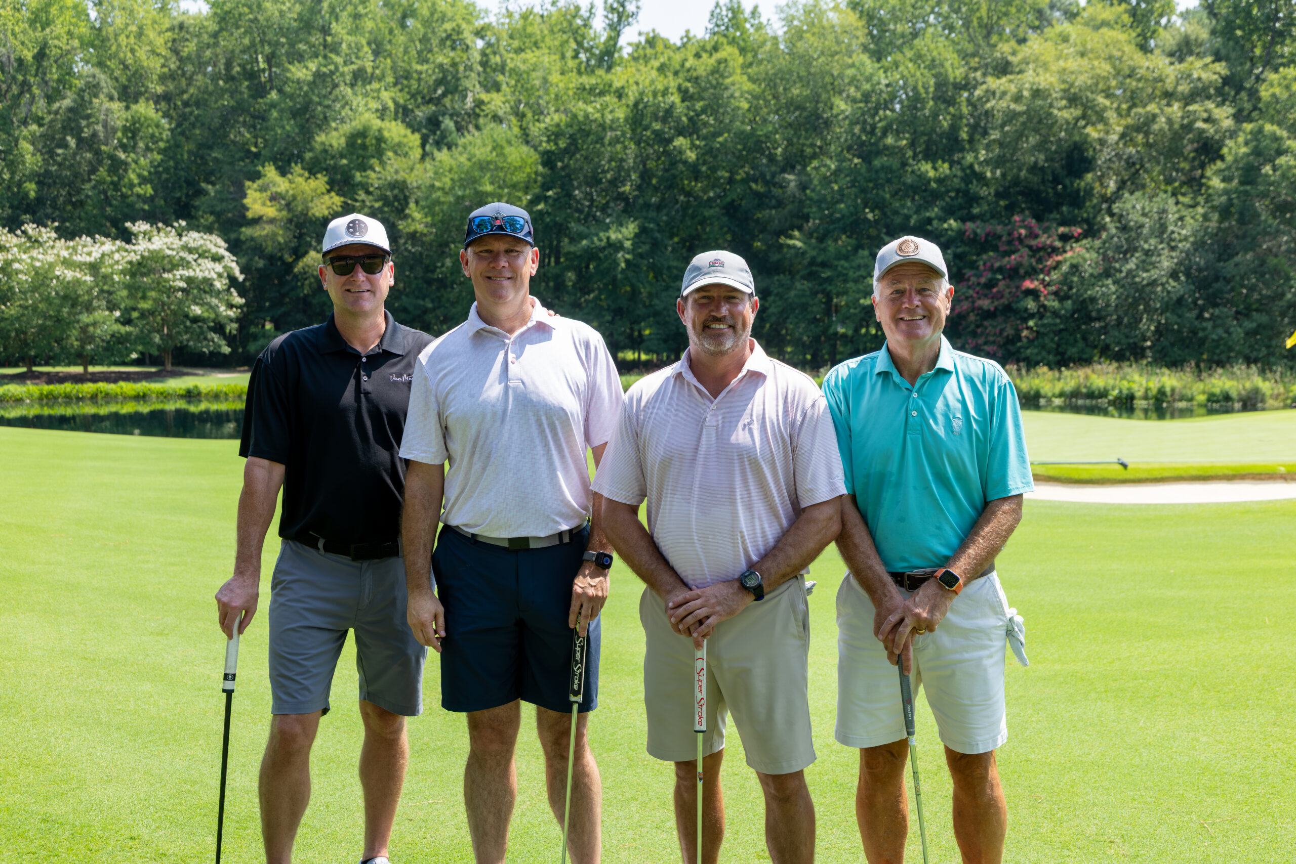 Four men wearing golf attire and caps stand together on a green lawn with clubs.
