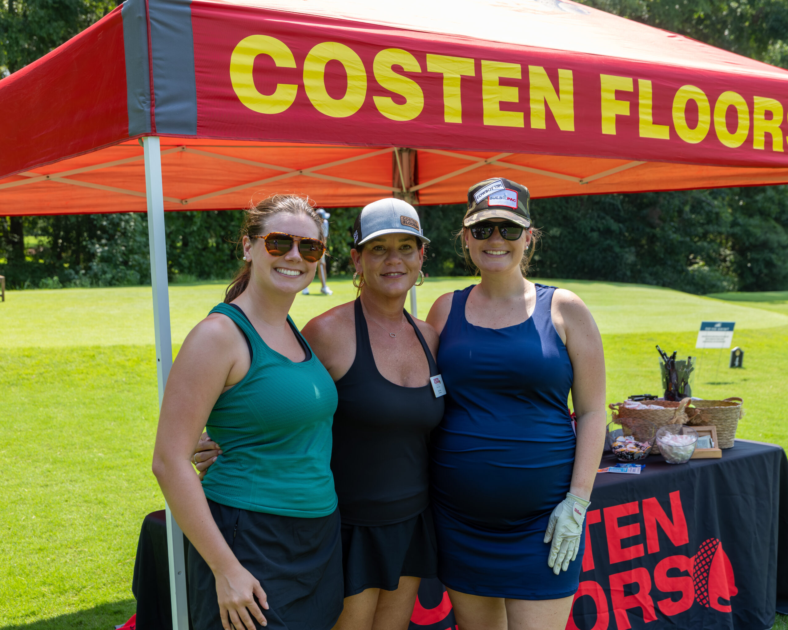 Three women standing together smiling under an orange and yellow banner for Costen Floors.