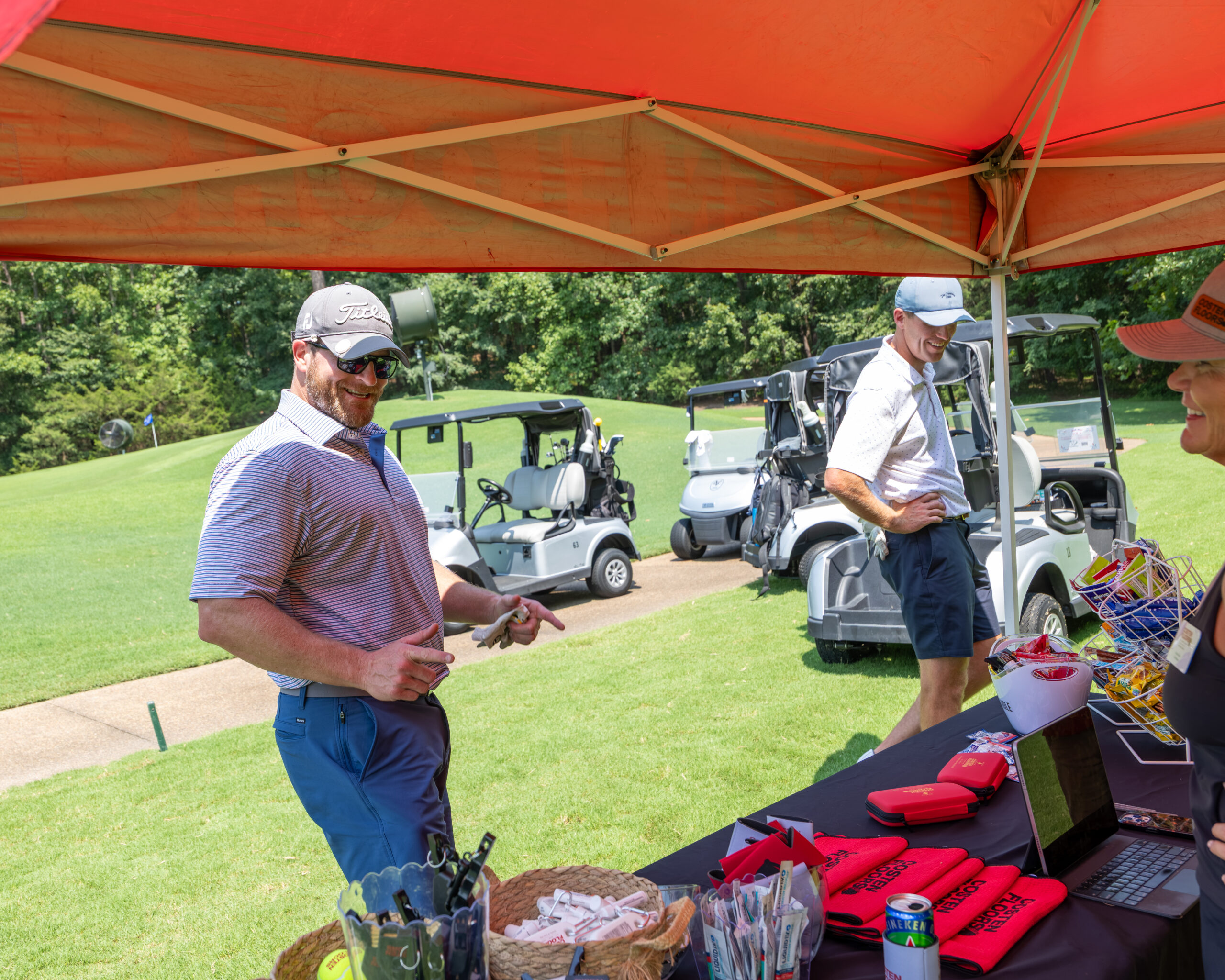 Two men and part of a woman under an orange tent at a golf tournament.
